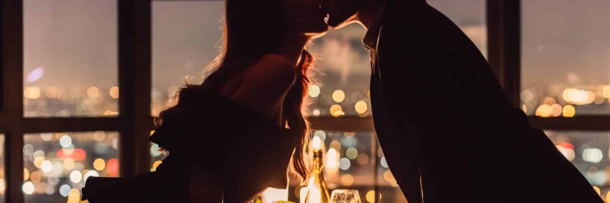 Loving couple kissing on panoramic window background overlooking nigth city. Romantic place for romantic evening concept